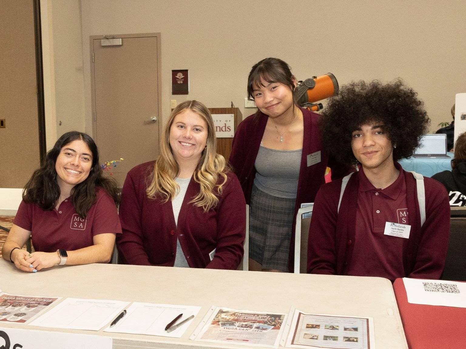Four students smiling behind a table while representing the Maroon & Grey Student Ambassadors.