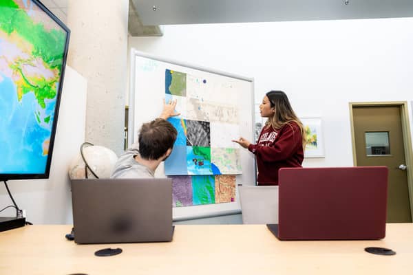 students in conference room with laptops open while looking at printed maps attached to whiteboard