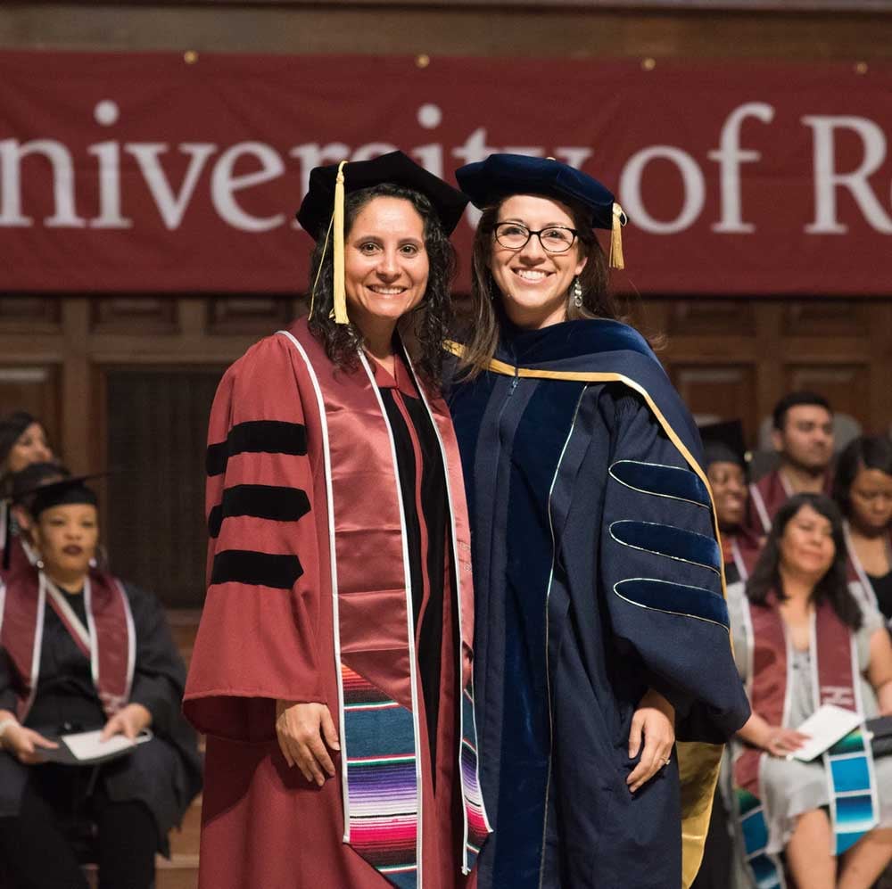 Two women pose on stage together wearing doctoral regalia
