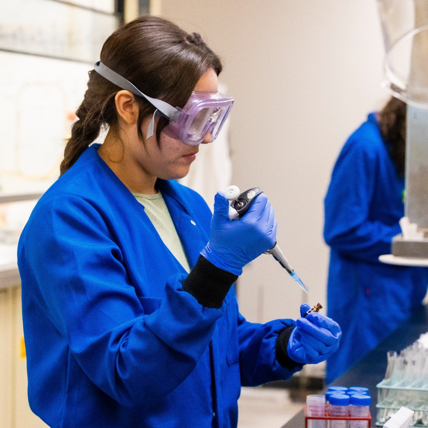 a woman wearing goggles and blue coat holding a pipette
