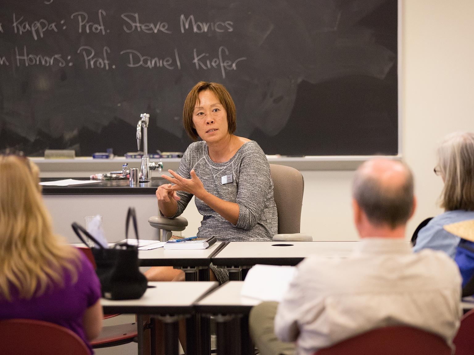 a woman sitting at a desk in front of a chalkboard
