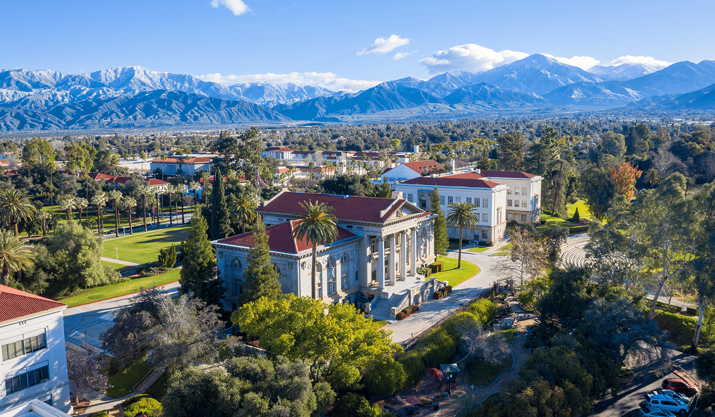 a large white building with a red roof surrounded by trees and mountains