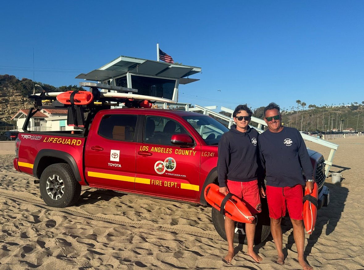 a couple of men standing next to a red truck with lifeguard equipment
