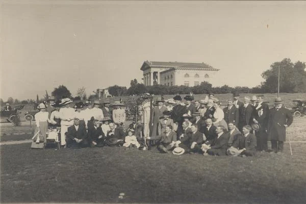 Group of people post with a large building in the background, sepia image, early 20th century