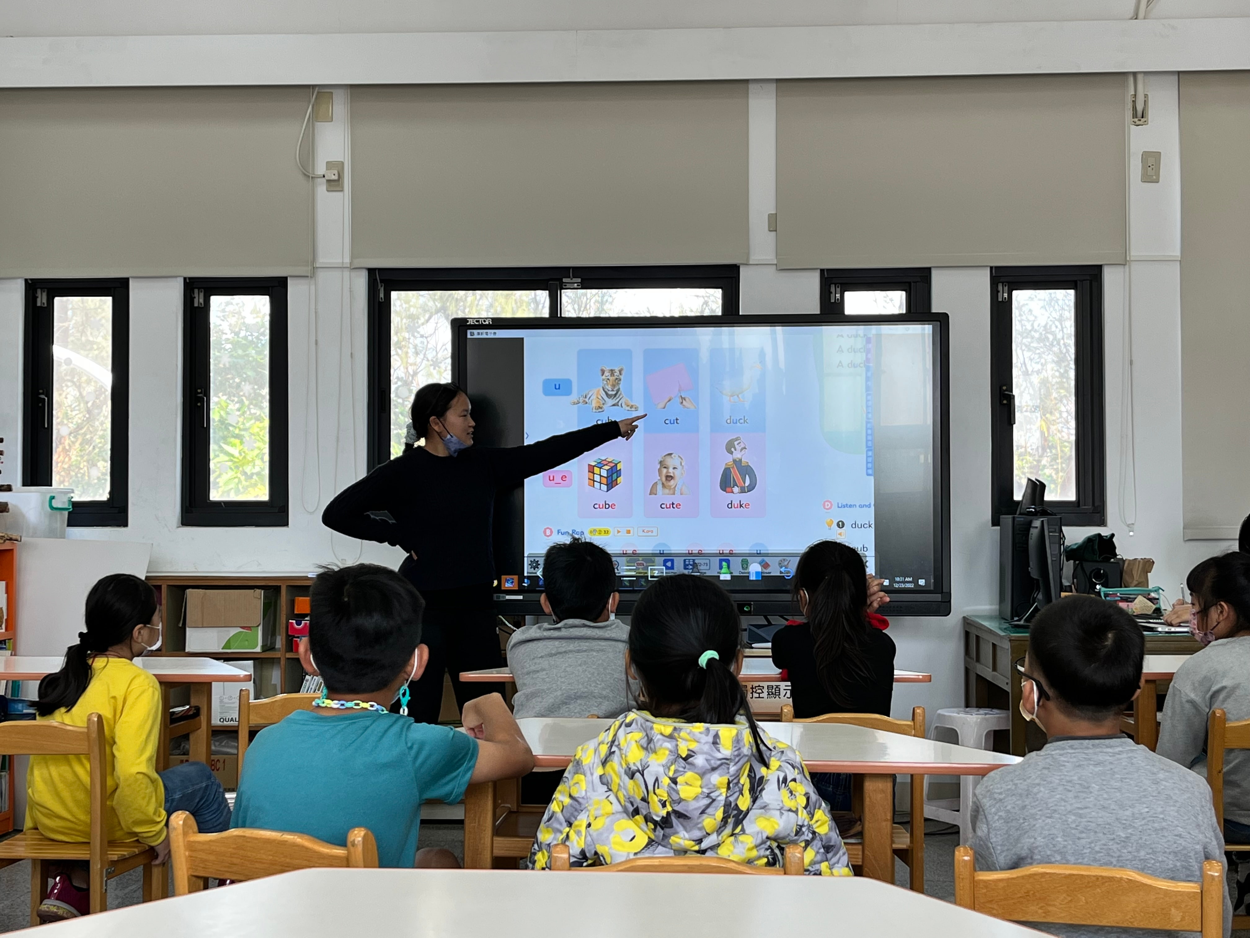 a woman standing in front of a large screen with a group of kids sitting at desks