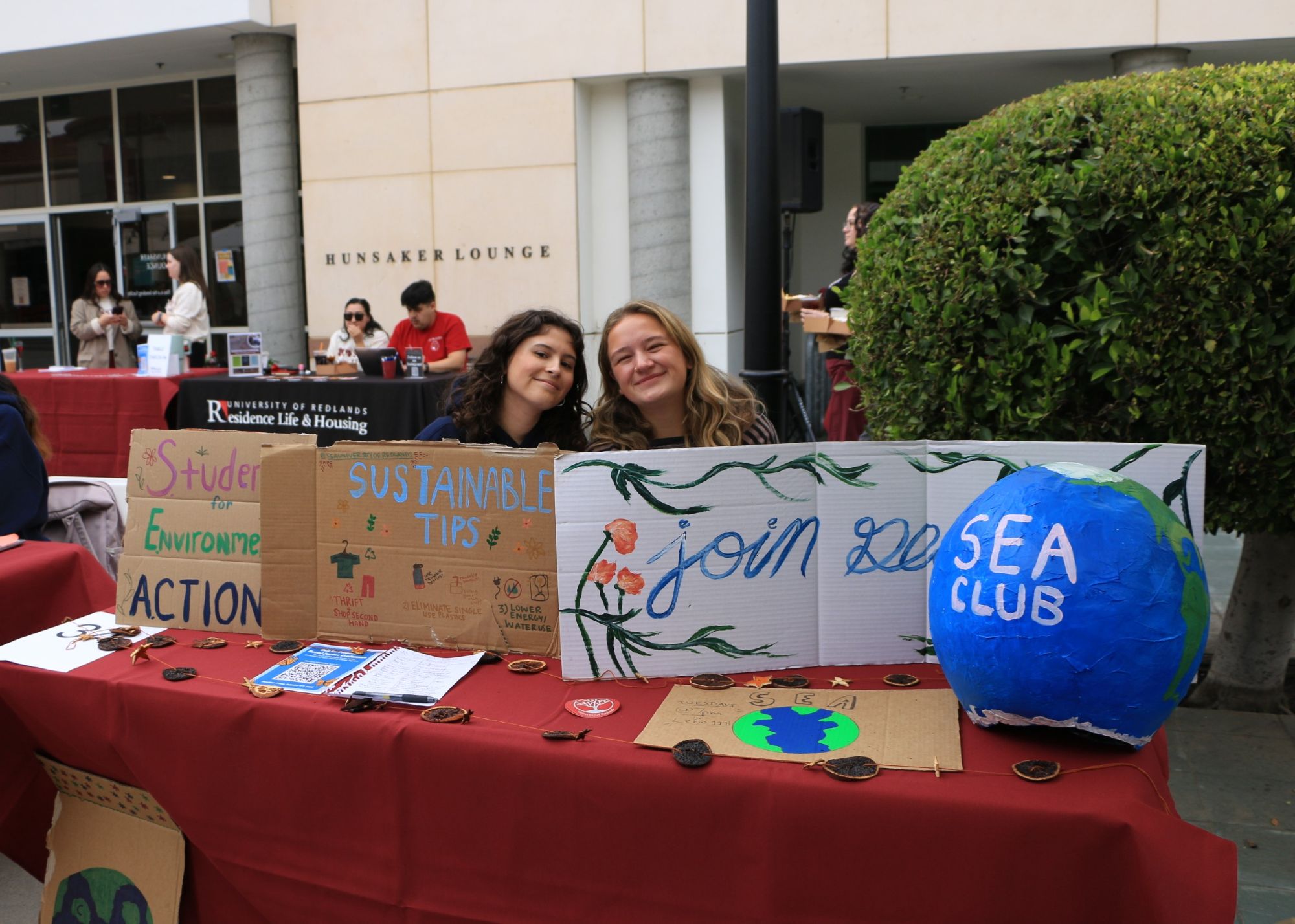 Two students sit behind a table with a large globe and handmade signs promoting the club and sustainability tips.
